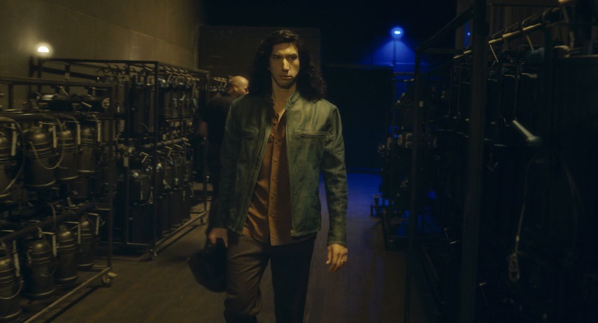 Adam Driver's character in a green leather jacket walking backstage.